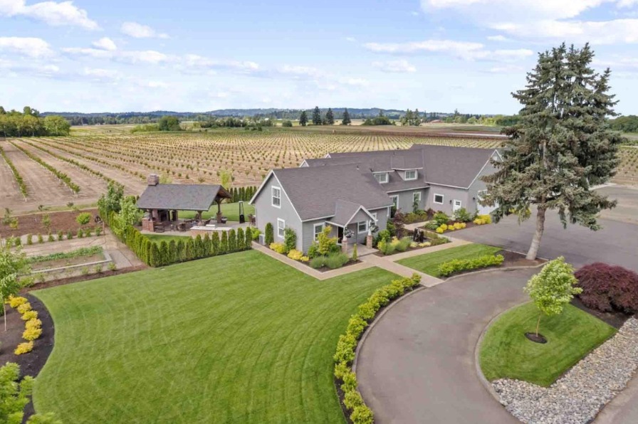 Aerial view of a charming gray house surrounded by lush green lawns and neatly arranged flower beds, with expansive orchards in the background.