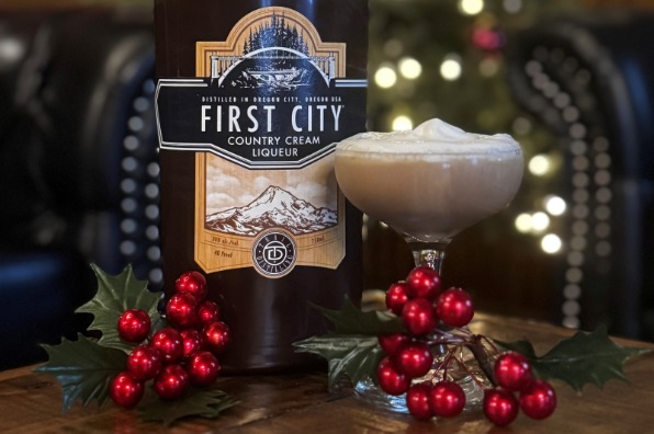 A bottle of First City Country Cream Liqueur sits on a wooden table, accompanied by a cocktail glass topped with whipped cream and festive decorations. A softly lit Christmas tree is in the background