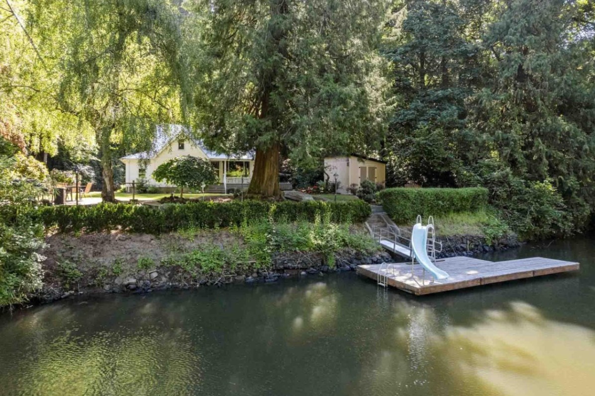 A serene riverside scene featuring a house surrounded by lush greenery, with a wooden dock and a slide leading into the water.