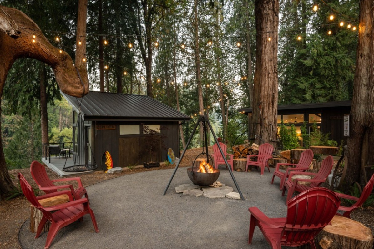 Cozy outdoor gathering space with red chairs around a fire pit, surrounded by tall trees and string lights, creating a warm atmosphere.