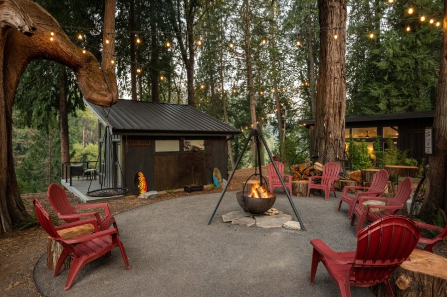 Cozy outdoor gathering space with red chairs around a fire pit, surrounded by tall trees and string lights, creating a warm atmosphere.