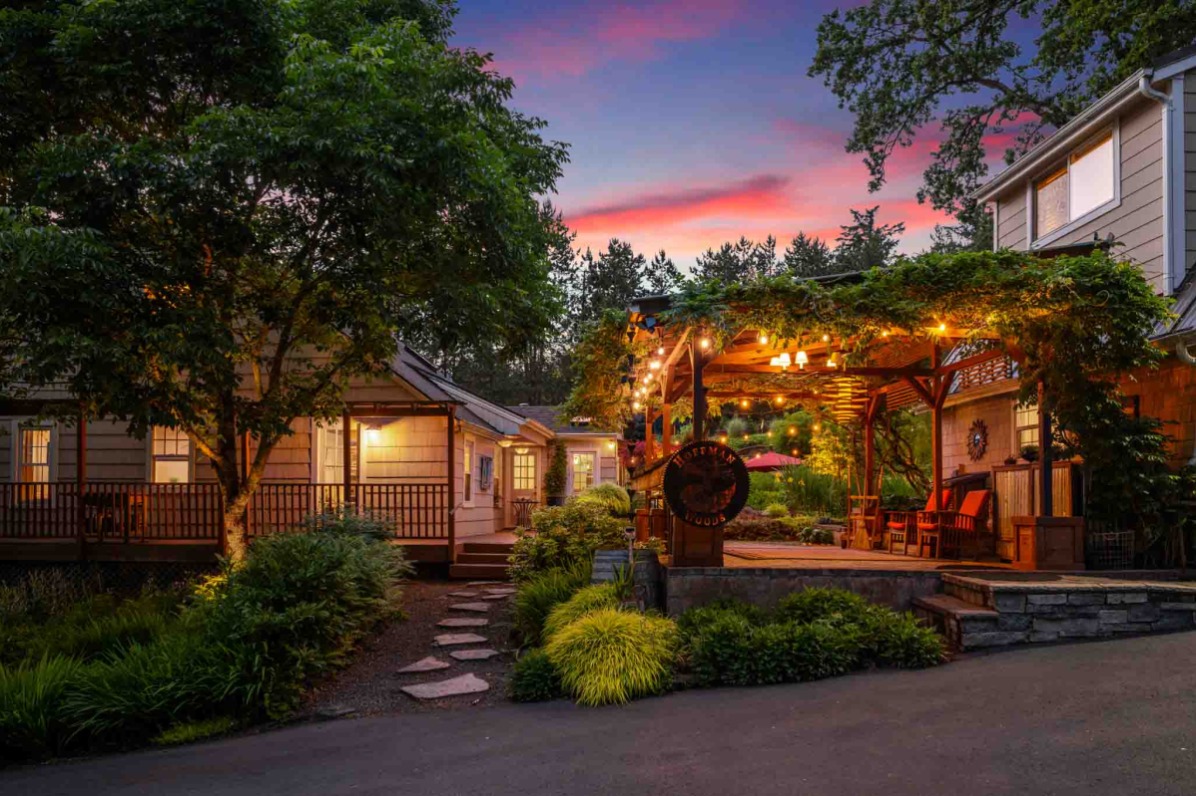 Charming outdoor seating area with string lights, surrounded by lush greenery and cozy cottages under a colorful sunset sky.