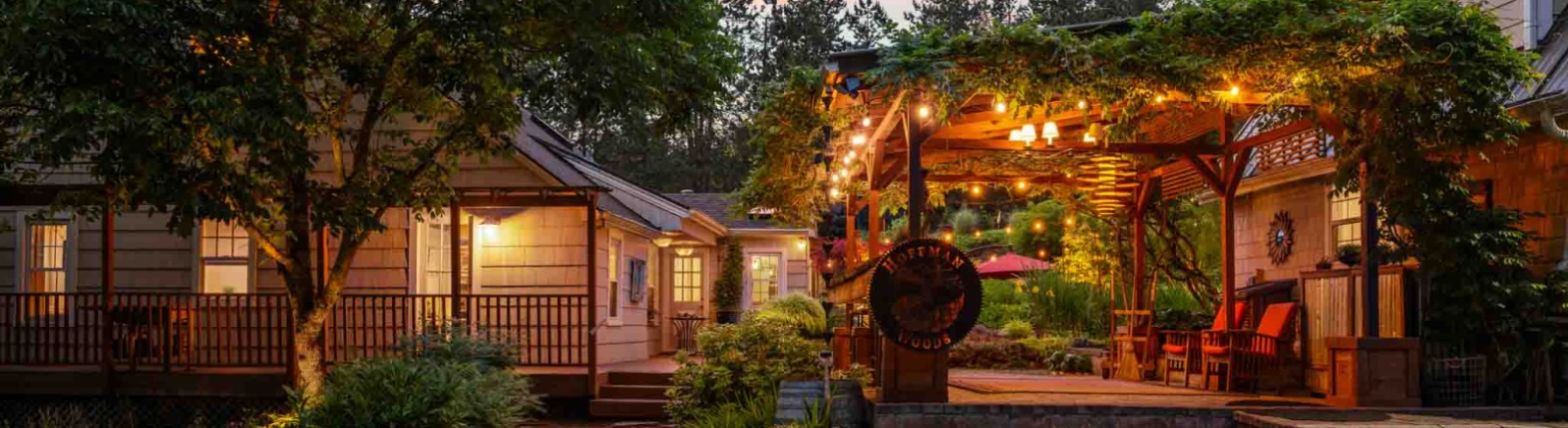 Charming outdoor seating area with string lights, surrounded by lush greenery and cozy cottages under a colorful sunset sky.