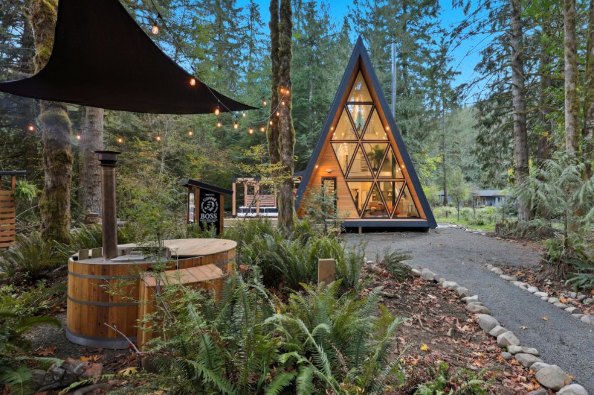 A modern A-frame cabin nestled among tall trees, surrounded by lush greenery. In the foreground is a wood burning hot tub. 
