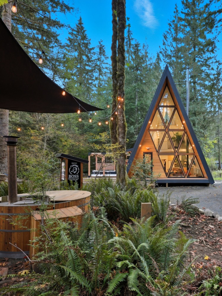 A modern A-frame cabin nestled among tall trees, surrounded by lush greenery. In the foreground is a wood burning hot tub. 