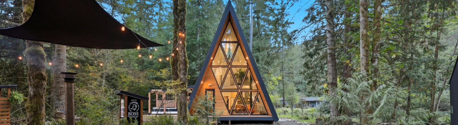 A modern A-frame cabin nestled among tall trees, surrounded by lush greenery. In the foreground is a wood burning hot tub. 