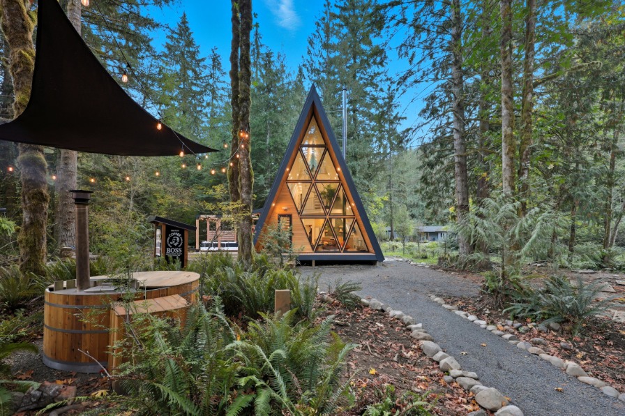 A modern A-frame cabin nestled among tall trees, surrounded by lush greenery. In the foreground is a wood burning hot tub. 