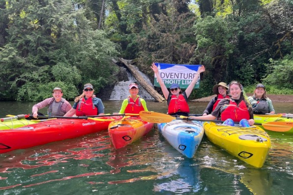A group of kayakers pose for a photo in front of a small waterfall while holding an Adventure Without Limits banner