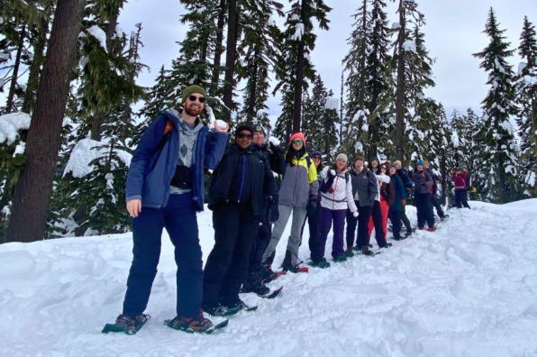 A large group of snowshoers stand in a line holding snowballs in a snowy forest