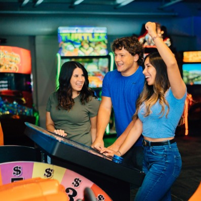 Three friends enjoy a vibrant arcade, engaging with a large game console. Colorful machines and neon lights create an energetic atmosphere.