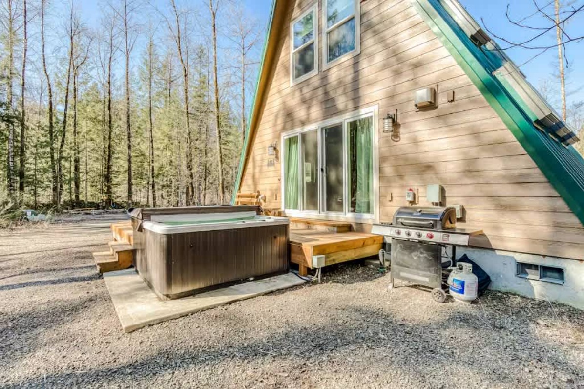 A cozy A-frame cabin surrounded by tall trees, featuring a hot tub and a grill on a wooden deck, inviting relaxation in nature.