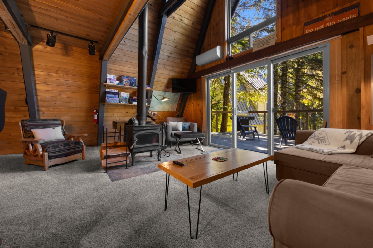 Cozy cabin interior featuring wooden walls, a fireplace, and large windows overlooking a forested deck. 