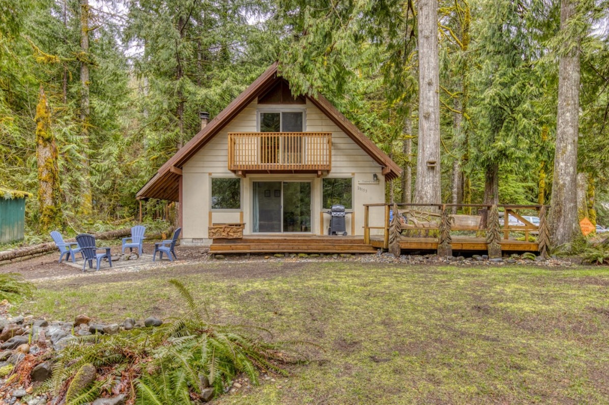 A cozy A-frame cabin surrounded by tall evergreen trees, featuring a wooden deck and outdoor seating area on a grassy lawn.