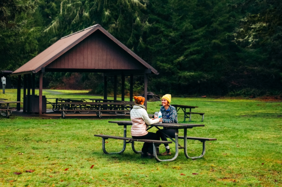 Two people in beanie and jackets sit facing each other at a picnic table in a green space with a covered seating area and trees in the background