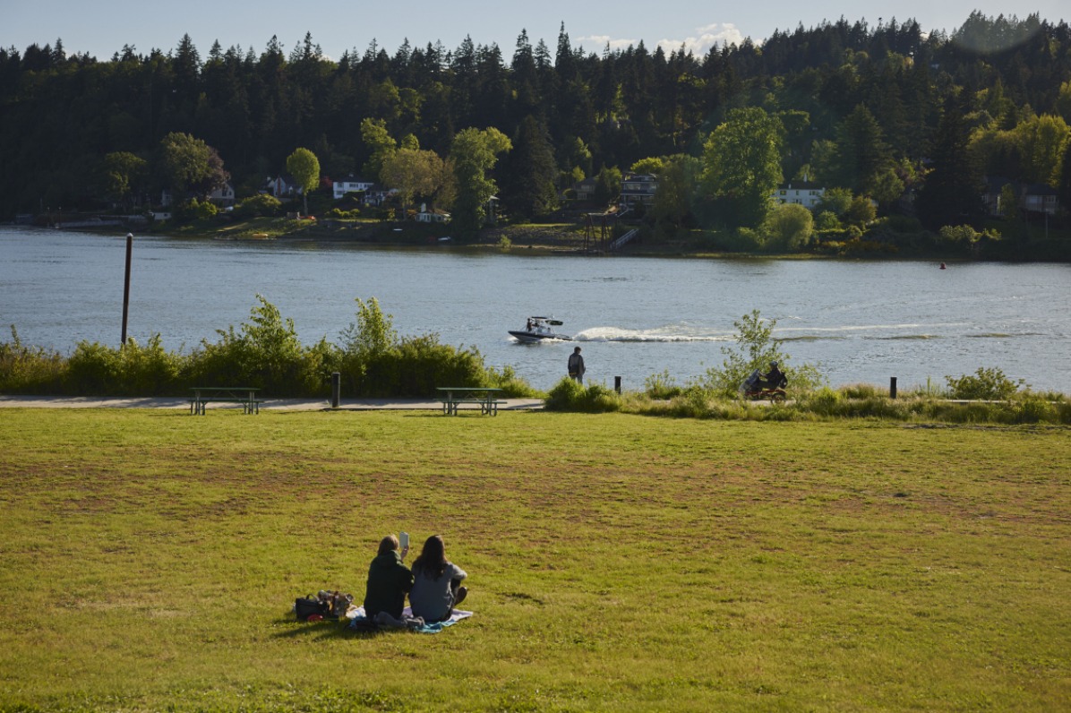 A couple sits on a grassy area looking at a river where there's a boat gliding across the water.