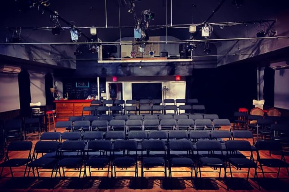 Empty theater space with rows of black chairs facing a stage, illuminated by overhead lights. A bar area is visible in the background.
