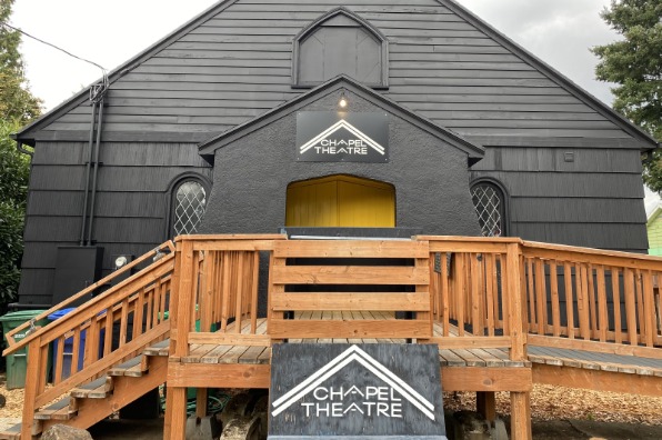 Black building with yellow door and cedar porch with ramp and stairs. Sign reads Chapel Theater.