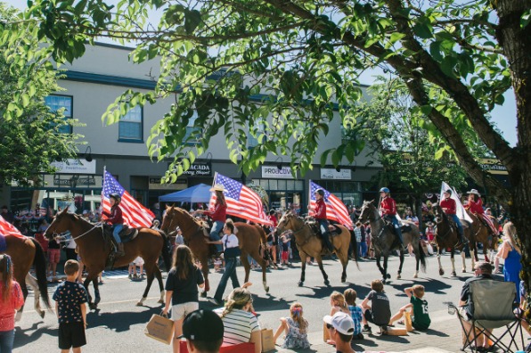 A lively parade scene with riders on horseback carrying American flags, surrounded by spectators and storefronts under a clear sky.