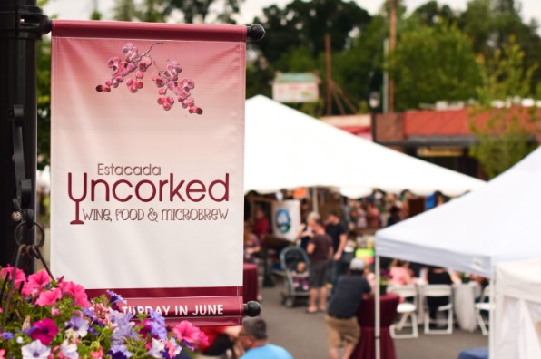 Banner for "Estacada Uncorked" event featuring wine, food, and microbrew. Background shows a bustling outdoor festival with tents and attendees.