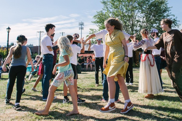 A lively outdoor gathering with people dancing on grass, surrounded by trees and a clear blue sky. Various ages participate in the celebration.