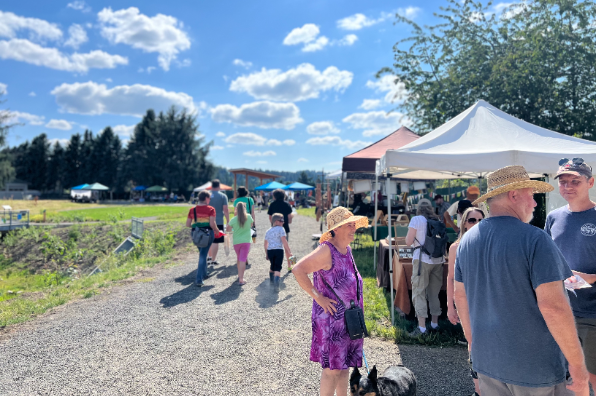 A vibrant outdoor market scene with people browsing stalls under a clear blue sky, surrounded by greenery and distant trees.