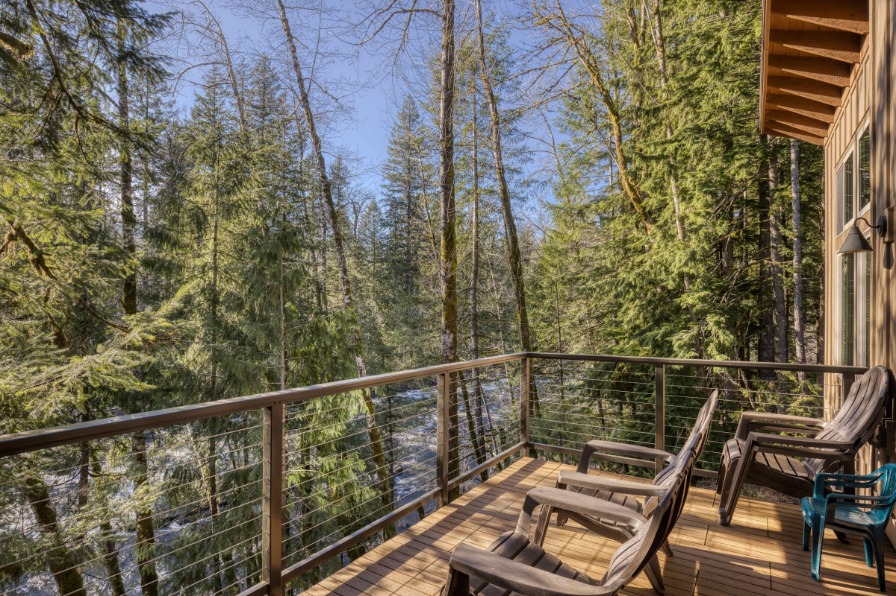 A wooden balcony overlooks a lush forest with tall trees, featuring two black lounge chairs and a view of a flowing river below.