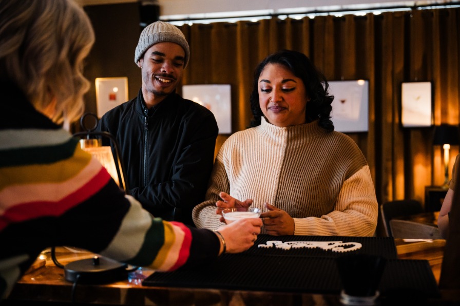 A cozy bar scene featuring two people engaged in conversation while a bartender prepares a drink. Warm lighting and art on the walls enhance the atmosphere.