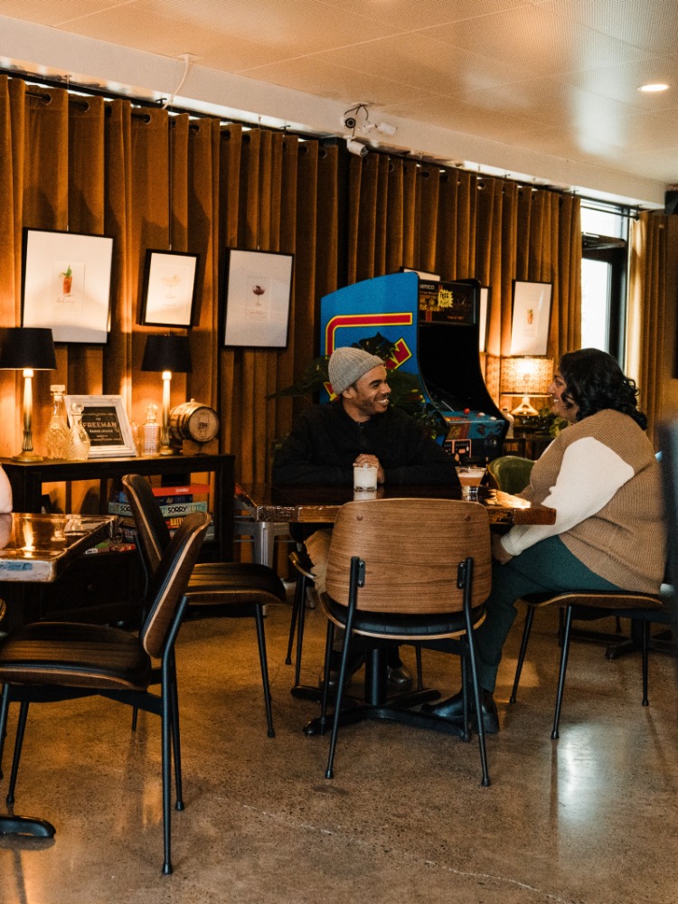 A cozy bar scene featuring people engaged in conversation. Warm lighting and art on the walls enhance the atmosphere.