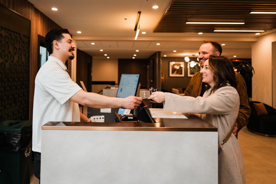 Hotel front staff assists a couple at the front desk, handing them a card. Modern decor and warm lighting create a welcoming atmosphere.