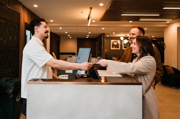 Hotel front staff assists a couple at the front desk, handing them a card. Modern decor and warm lighting create a welcoming atmosphere.