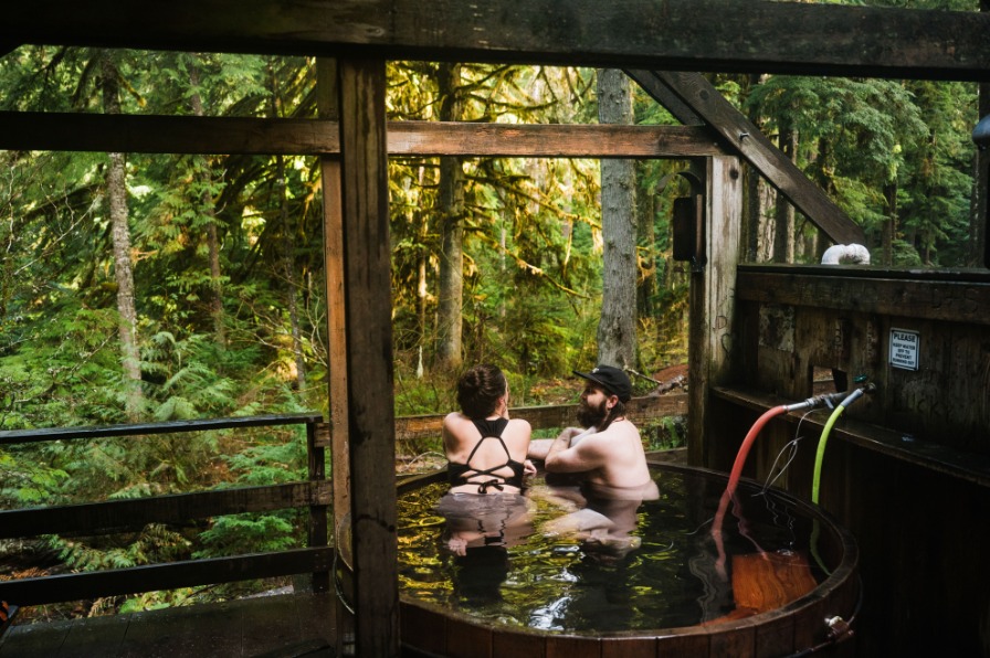 A wooden hot tub surrounded by lush green forest, with two people relaxing inside. Sunlight filters through the trees, creating a serene atmosphere.