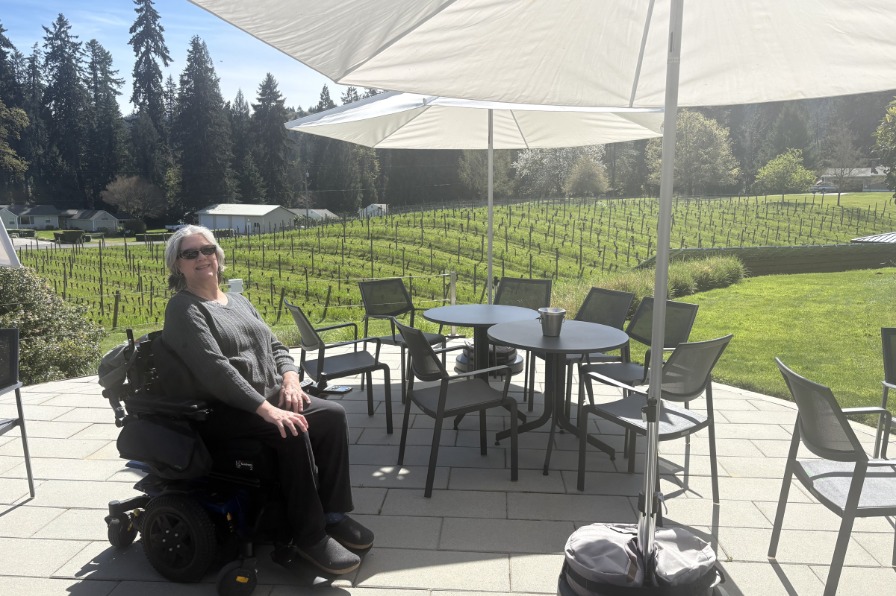 Twill Cellars A person in a wheel chair sits on a winery patio with vines behind.