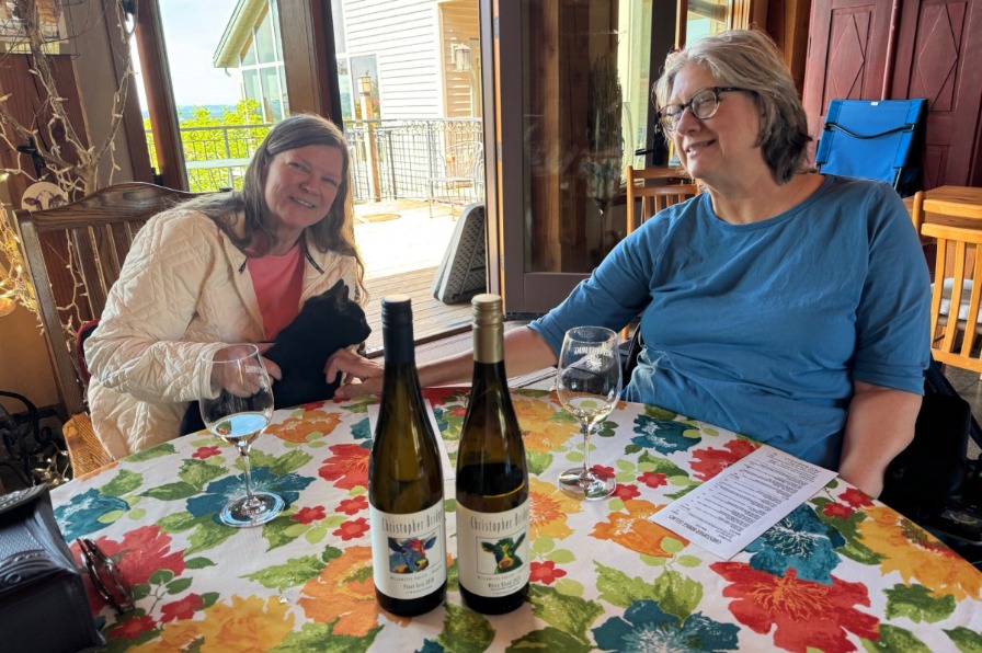 Christopher Bridge Cellars Two people pet a black cat with wine bottles on a table.