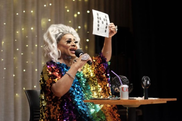Drag Queen Poison Waters is dressed in a rainbow colored shimmery dress and holds up an example bingo card.