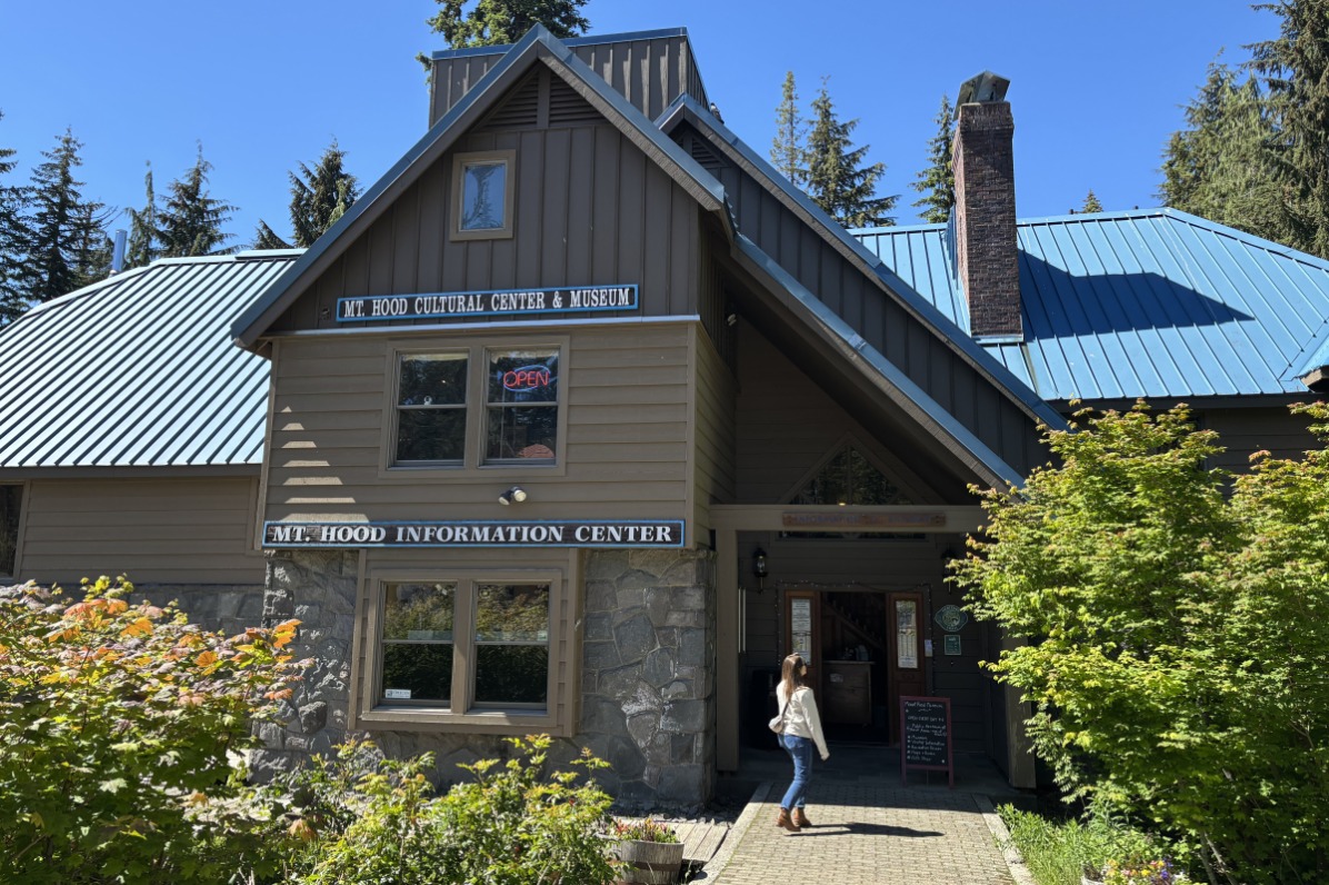 A visitor approaches the entrance to Mt. Hood Cultural Center & Museum, a rustic building with a blue roof and stone accents, surrounded by greenery.