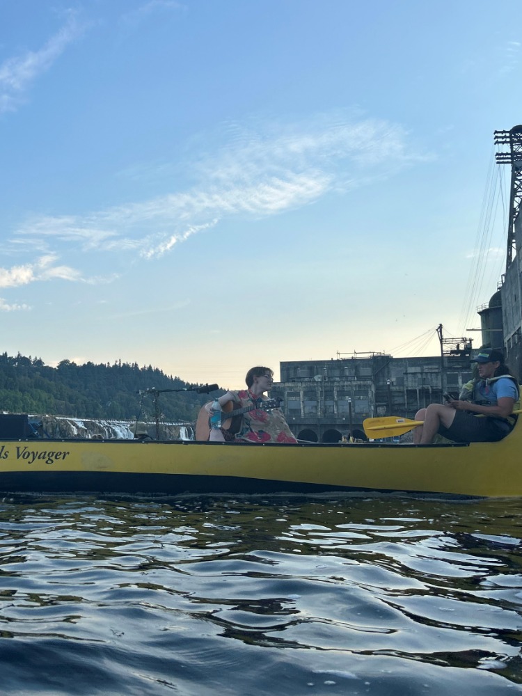 A musician sings any plays guitar in a long yellow canoe