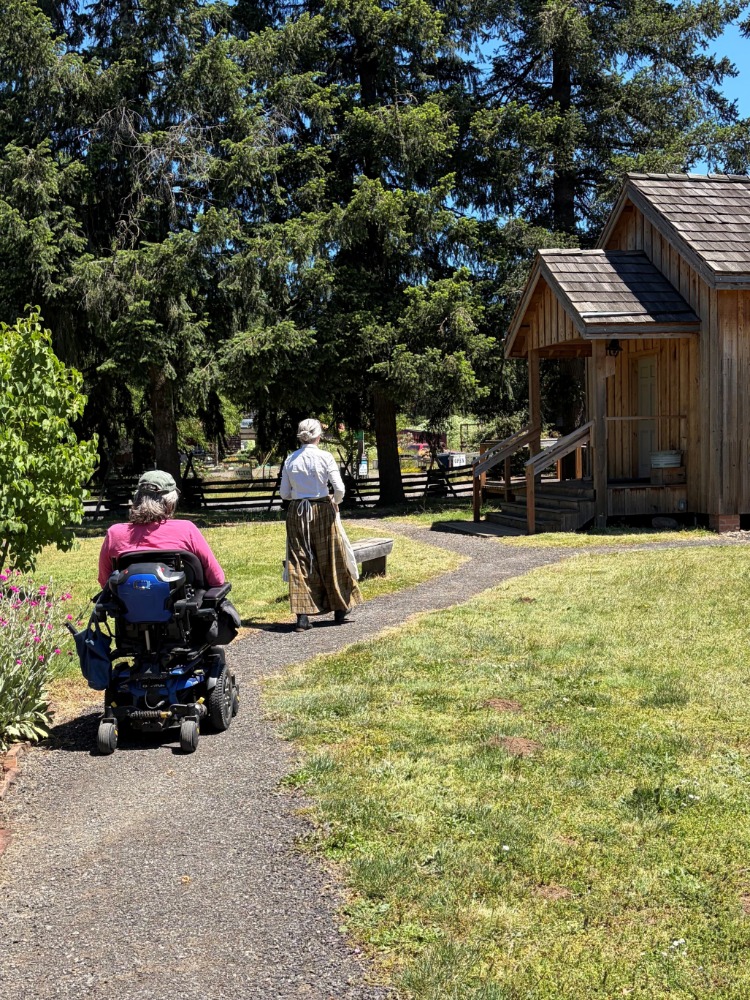A person in a wheelchair and a person in pioneer garb on a paved path near historic buildings