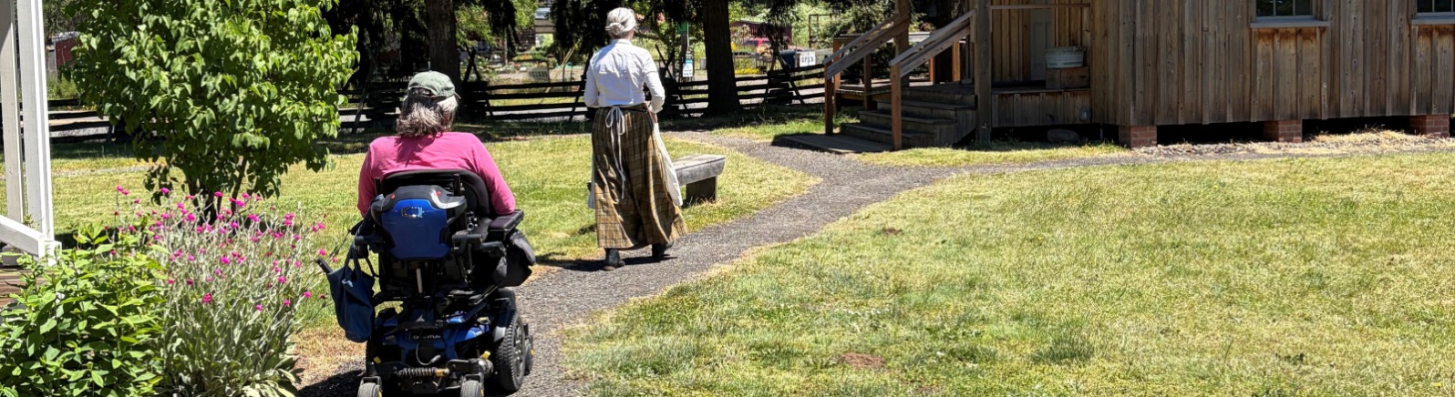 A person in a wheelchair and a person in pioneer garb on a paved path near historic buildings