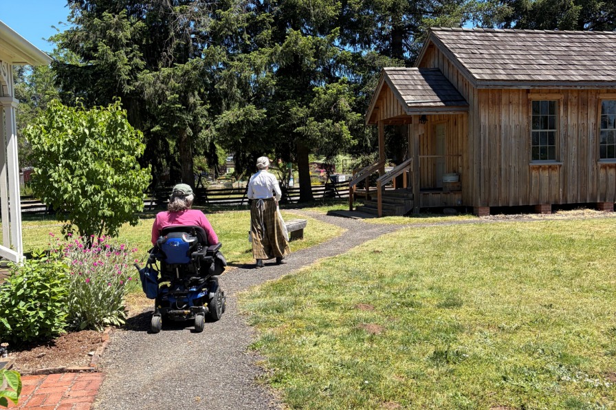 A person in a wheelchair and a person in pioneer garb on a paved path near historic buildings