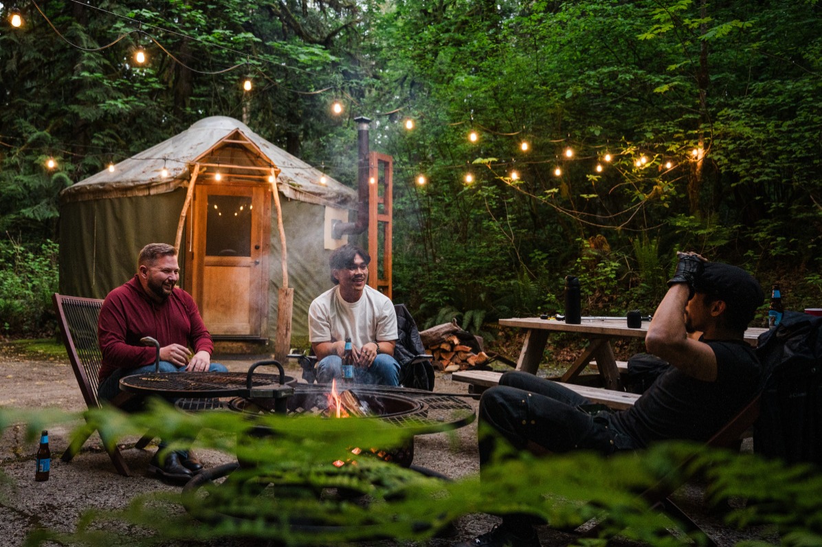 People sit around a campfire with a yurt behind them and green trees