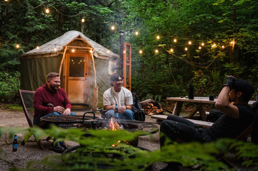 People sit around a campfire with a yurt behind them and green trees