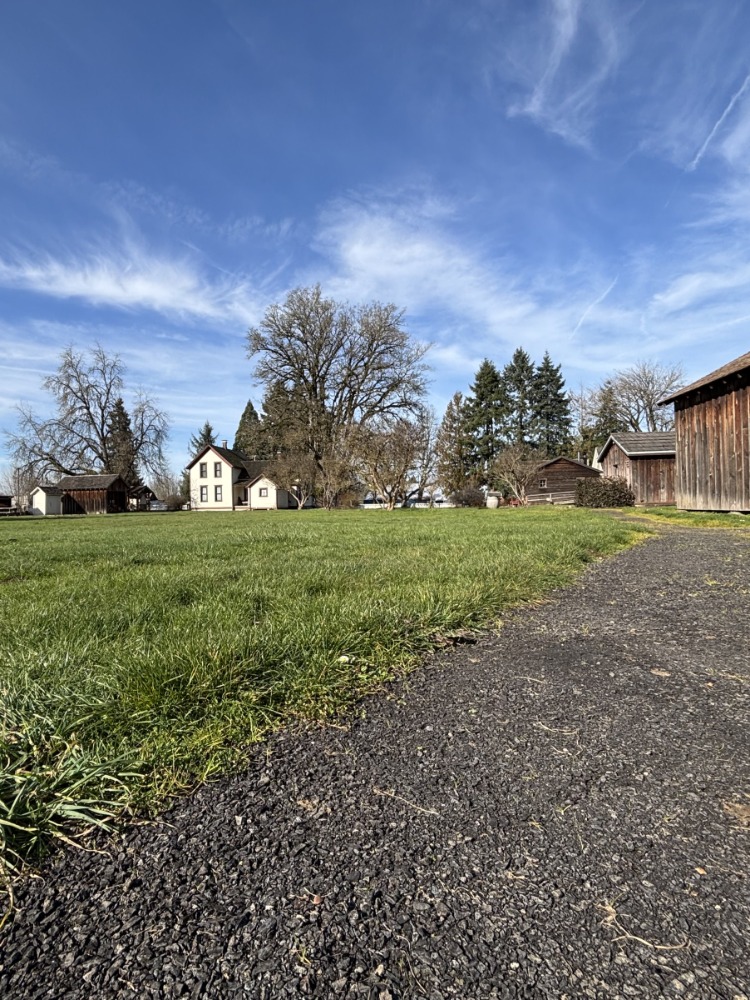 An accessible packed-gravel path leads to a cluster of rustic buildings under a clear blue sky, surrounded by trees.