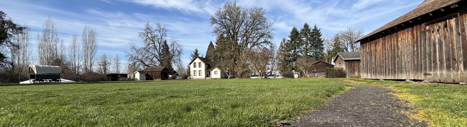 An accessible packed-gravel path leads to a cluster of rustic buildings under a clear blue sky, surrounded by trees.