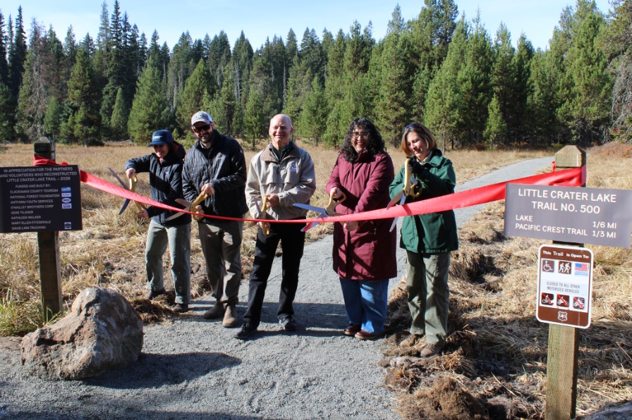 A group of people cut a red ribbon on a grey gravel trail with green trees behind.