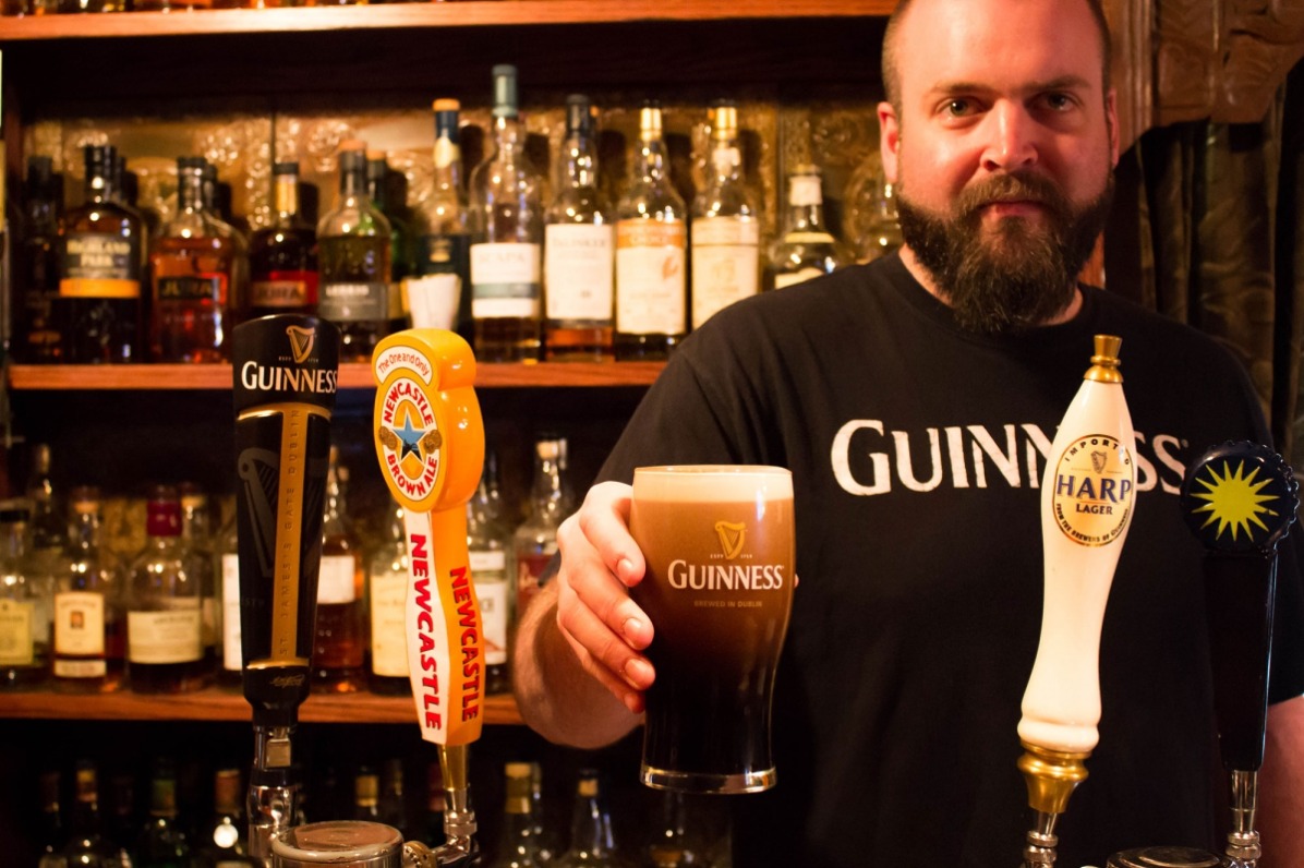 Highland Still House back bar of liquor bottles and bearded bartender in Guinness shirt holding out a glass of Guinness Ale