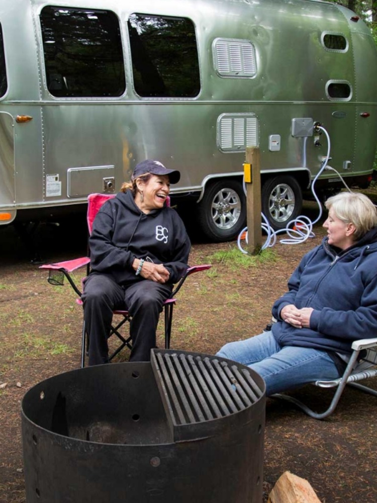 Two women sit in camp chairs enjoying the outdoors with their Airstream trailer and bicycle behind them in their camping site