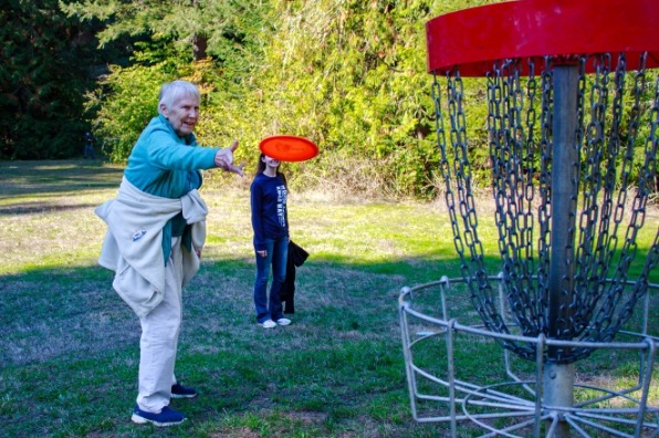 Senior lady tosses red frisbee and teenage girl looks on as they play disc golf at a park in Oregon's Mt. Hood Territory