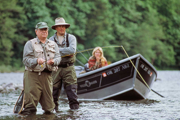 Two people in waders and fishing vests are standing in Sandy River while a third person remains in drift boat