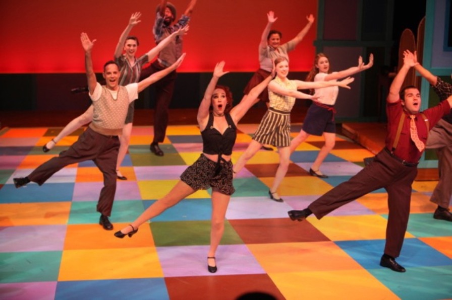 Enthusiastic troupe of theater performers belt out a song while dancing on a stage of multi-color squares